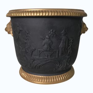 May include: Black planter with a gold rim and base, featuring a detailed relief scene. The planter has two gold lion head handles. The relief depicts figures in a landscape. A decorative piece for home or garden.