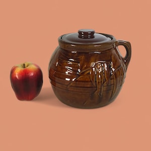 Puede incluir: Un tarro de cerámica marrón con tapa y asa, junto a una manzana roja. El tarro tiene un acabado brillante y un diseño texturizado. La manzana es roja con acentos amarillos.