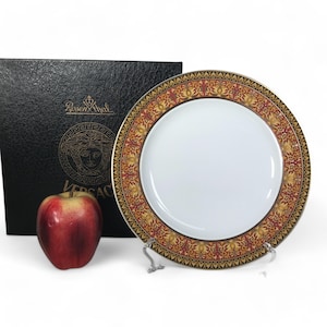 Pode incluir: Um prato de jantar de porcelana branca com uma borda estampada em ouro e vermelho. O prato está em um suporte e tem o texto "Rosenthal Versace" na caixa.
