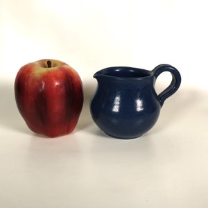 May include: A small, dark blue ceramic creamer or pitcher with a rounded body and a handle. The creamer is sitting on a white surface.