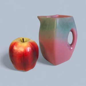 May include: A red and yellow apple next to a ceramic pitcher. The pitcher has a handle and a spout, with a color gradient from green to pink. The apple is shiny and appears to be ripe.