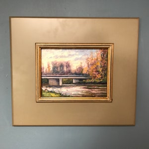 Pode incluir: Uma pintura a óleo emoldurada que retrata uma ponte sobre um rio com árvores de cada lado. O céu é azul claro com nuvens. As árvores são uma mistura de verde e marrom.