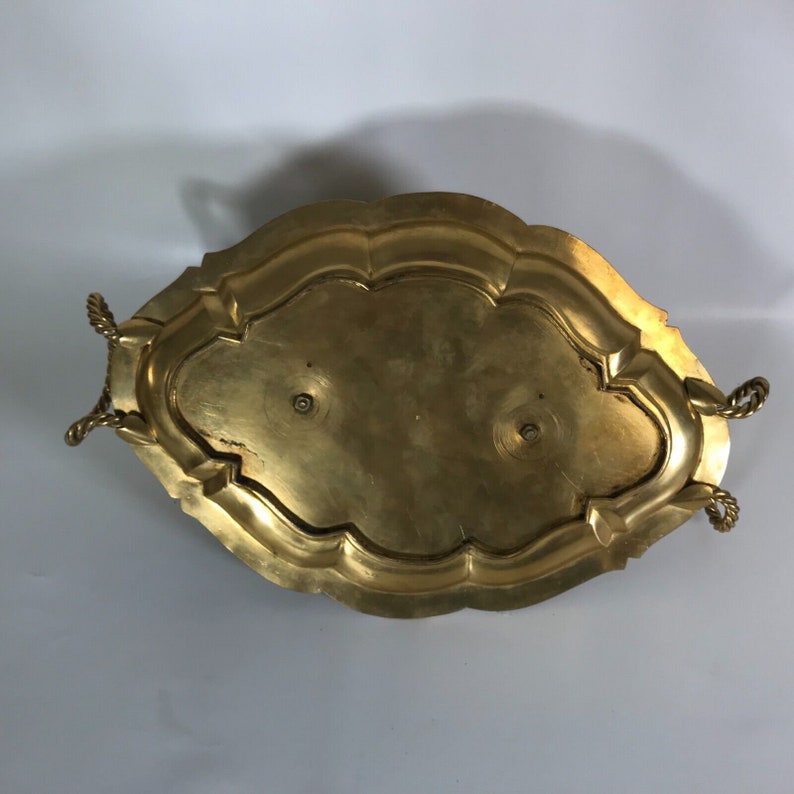 May include: A gold-toned metal serving tray with a rope-like handle on each side. The tray has a scalloped edge and a raised center.