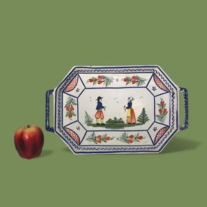 Könnte beinhalten: Ein rechteckiges Serviertablett aus Keramik mit blauen Griffen und einem dekorativen Rand. Das Tablett zeigt eine handgemalte Szene von zwei Figuren in traditioneller Kleidung, umgeben von floralen Motiven. Links liegt ein roter Apfel. Das Tablett ist ca. 30 cm lang.