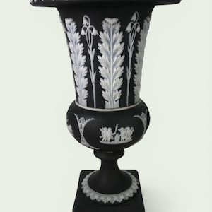 May include: Black and white decorative urn with a square base. The urn features detailed white relief designs of leaves, flowers, and classical figures. The top rim and base have ornate leaf patterns. A classic home decor item.
