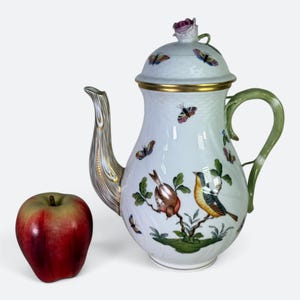 May include: A white porcelain teapot featuring hand-painted colorful birds, butterflies, and green foliage. The teapot has a gold-trimmed spout, a green handle, and a lid topped with a sculpted pink rose. A red and yellow apple sits beside it.