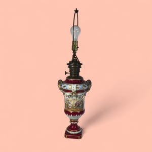 May include: An antique table lamp with a burgundy and white porcelain base, featuring a detailed painted scene. The lamp has a brass fixture and a light bulb at the top. The lamp's design includes ornate handles and decorative accents.