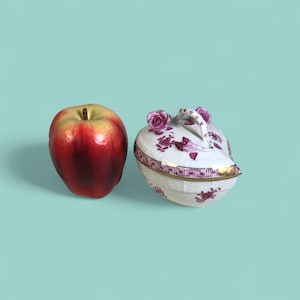 Op de afbeelding: Een rood-gele appel naast een witte porseleinen sieradendoos met een deksel. De doos is versierd met paarse bloempatronen en gouden randen. Het deksel heeft een handvat en twee roosvormige accenten.
