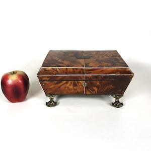May include: A small wooden box with a brown and white marbled finish. The box has a hinged lid and a brass lock. It sits on four decorative brass feet.