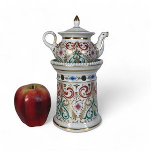 Puede incluir: Una tetera de porcelana blanca con diseños florales dorados y rojos se asienta sobre un soporte a juego. La tetera tiene una tapa y un pico. El soporte tiene una base redonda y un cuerpo cilíndrico.