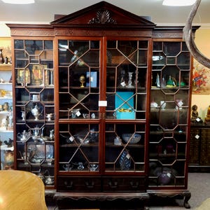 May include: A large, dark wood display cabinet with glass doors and geometric patterns. The cabinet is filled with various decorative items, including silver tea sets, figurines, and glassware. The cabinet has a decorative top and sits on a carved base.