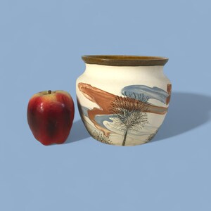 May include: A ceramic vase with a cream-colored base, featuring a brown and blue abstract design with a tree illustration. A red apple sits to the left of the vase. The vase has a brown rim.