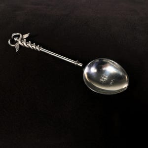 Può includere: Un cucchiaio d'argento con un manico attorcigliato e un design decorativo di uccello nella parte superiore. Il cucchiaio ha una ciotola arrotondata ed è inciso con il testo "Made in England".