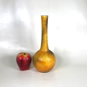 May include: A tall, slender, orange and yellow vase with a bulbous base and a long neck. The vase has a textured surface and a dark brown pattern near the top. A red apple sits to the left of the vase.