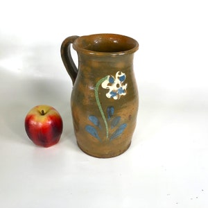 May include: A brown ceramic jug with a white flower and blue leaves painted on the side. The jug has a handle and a rounded body.