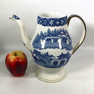 Puede incluir: Una tetera de porcelana azul y blanca con asa dorada. La tetera tiene un diseño floral y una escena inspirada en la arquitectura china.
