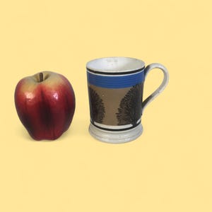 Puede incluir: Una taza de cerámica con interior blanco, una franja azul y una banda marrón con un diseño de árbol negro. La taza tiene un asa y una base blancas. Una manzana roja está al lado de la taza.