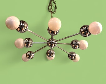 May include: A mid-century modern chandelier with a starburst design. The fixture features multiple chrome arms extending from a central sphere, each holding a white globe and a chrome sphere. The light hangs from a brown chain against a green background.