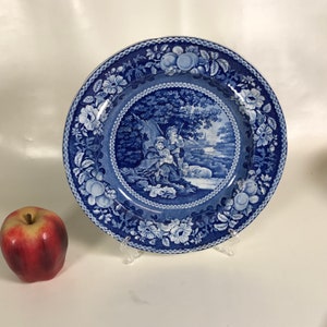 May include: A blue and white porcelain plate with a floral border and a central image of two children in a woodland setting. The plate is on a white surface.