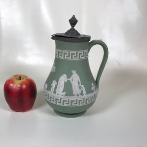 May include: A sage green ceramic pitcher with a lid and handle, featuring white relief figures and a Greek key border. A red apple sits to the left of the pitcher. The pitcher is approximately 10 inches tall.