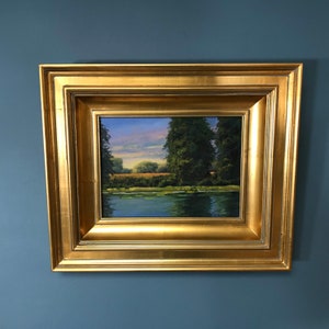 Pode incluir: Uma pintura de paisagem emoldurada que retrata uma cena de rio com árvores e uma margem gramada. A pintura está em uma moldura dourada com uma superfície texturizada.