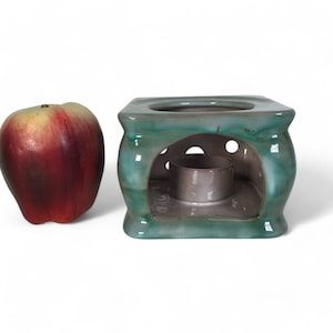 May include: A teal ceramic oil burner with a small, round, white ceramic dish inside. The burner has a square base and a rounded top.