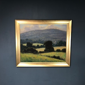 Pode incluir: Uma pintura de paisagem emoldurada em uma moldura dourada. A obra de arte retrata colinas verdes, árvores e uma montanha distante sob um céu nublado. A pintura está montada em uma parede azul escura.