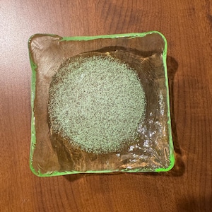 May include: A square glass dish with a light green rim and a central area filled with a light green granular substance. The dish is transparent, allowing the brown surface beneath to be visible.