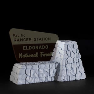 May include: A miniature model of a sign for the Pacific Ranger Station in Eldorado National Forest. The sign is brown and beige, with the text in white. The base is a light blue stone structure.