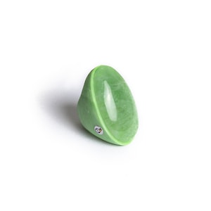 Pode incluir: Um anel em forma de taça verde-claro com um acabamento liso e polido e uma textura marmoreada. A banda apresenta uma pequena incrustação metálica circular de tom prateado na lateral, contrastando com as curvas suaves do material verde.
