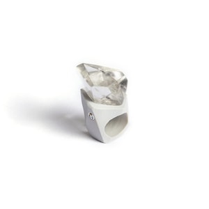 Pode incluir: Um anel moderno com um grande cristal claro e facetado, montado em uma banda geométrica cinza claro. O anel tem um design único e contemporâneo, adequado para moda ou ocasiões especiais.