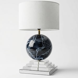 Puede incluir: Una lámpara de mesa decorativa con una pantalla blanca y una base de globo azul oscuro con vetas blancas. La base se asienta sobre una base rectangular clara y escalonada. La lámpara tiene un tallo color latón.