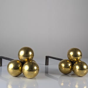 May include: A pair of fireplace andirons featuring a cluster of three polished brass spheres attached to a dark, L-shaped iron bar. The metallic gold spheres are arranged in a triangular formation, providing a reflective surface and a sturdy base.
