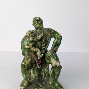 Puede incluir: Una escultura de cerámica verde de una figura sentada que sostiene una figura más pequeña. La escultura tiene una superficie rugosa y texturizada y está pintada en tonos verdes y marrones.