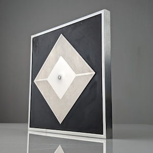 May include: Geometric wall art featuring a layered diamond design. The artwork has a black background with silver and white diamond shapes. It is encased in a silver metal frame, creating a modern, minimalist aesthetic.