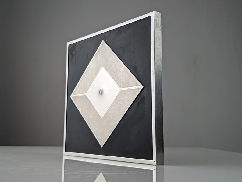 May include: Geometric wall art featuring a layered diamond design. The artwork has a black background with silver and white diamond shapes. It is encased in a silver metal frame, creating a modern, minimalist aesthetic.