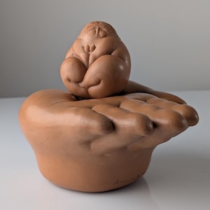 Puede incluir: Una escultura de cerámica color terracota que muestra una mano grande y abierta con los dedos extendidos, sosteniendo una pequeña figura sentada y redondeada con cabeza texturizada y cuerpo liso. La base incluye una firma en tinta negra.