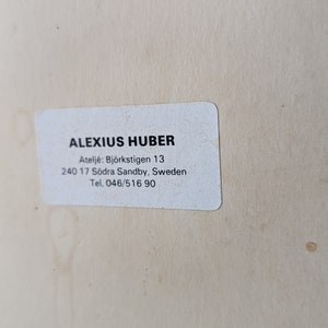 May include: A white rectangular sticker with the text "ALEXIUS HUBER" in black, along with an address and phone number. The sticker is on a light beige surface.