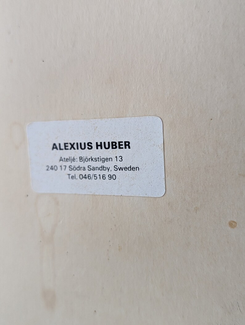 May include: A white rectangular sticker with the text "ALEXIUS HUBER" in black, along with an address and phone number. The sticker is on a light beige surface.