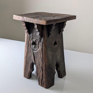 Könnte beinhalten: Ein rustikaler, quadratischer Holzschemel mit dunkelbraunem Finish. Der Hocker hat geschnitzte dekorative Elemente an den Beinen und Stützen und ist etwa 30 cm hoch. Die Oberseite ist quadratisch.