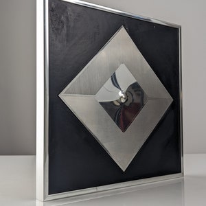 May include: A modern wall art piece featuring a black square background with a silver metal frame. The artwork has a layered design with a large silver diamond shape and a smaller, reflective diamond in the center.