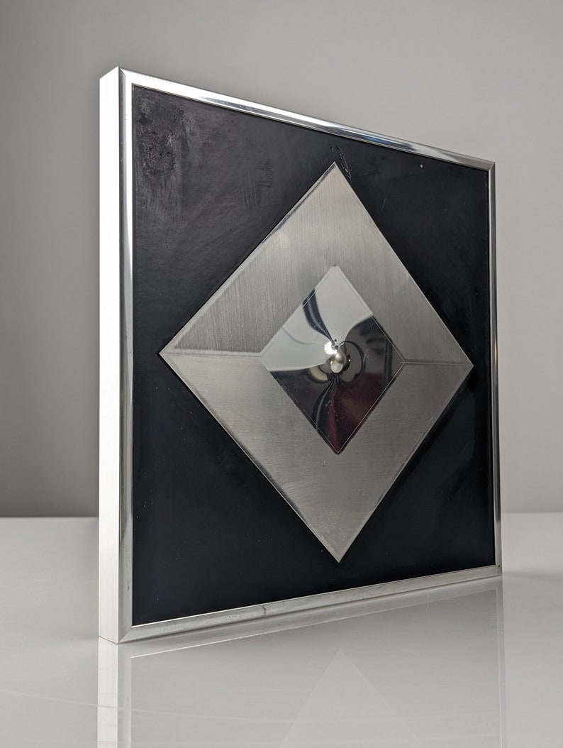 May include: A modern wall art piece featuring a black square background with a silver metal frame. The artwork has a layered design with a large silver diamond shape and a smaller, reflective diamond in the center.