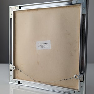 May include: The back of a framed artwork with a silver metal frame. The artwork's backing is a light tan color, with a small white label in the lower center. A wire is attached for hanging. The frame is square and has visible screws.