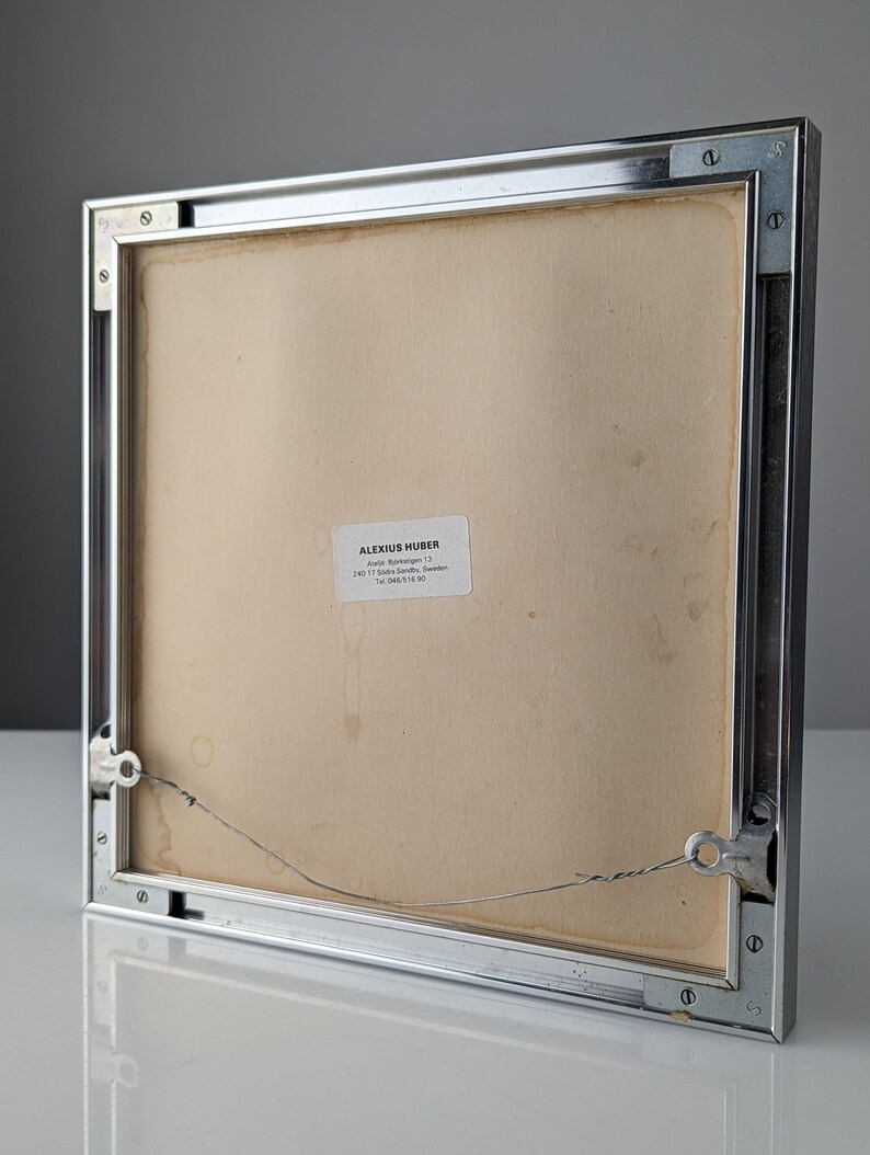 May include: The back of a framed artwork with a silver metal frame. The artwork's backing is a light tan color, with a small white label in the lower center. A wire is attached for hanging. The frame is square and has visible screws.