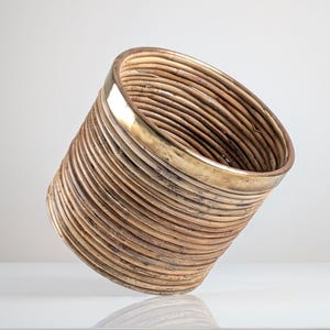 Puede incluir: Una cesta cilíndrica de ratán tejido con un borde color latón. La cesta está hecha de tiras de ratán marrón claro, muy enrolladas, creando una superficie texturizada. El diseño está ligeramente inclinado, mostrando la construcción en capas.