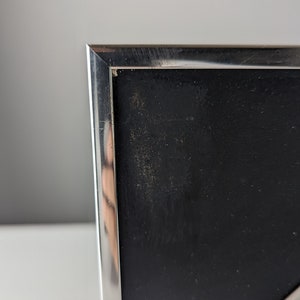 May include: Close-up of a silver metal picture frame with a black interior. The frame has a clean, modern design with a reflective surface. The black interior has a slightly textured appearance.