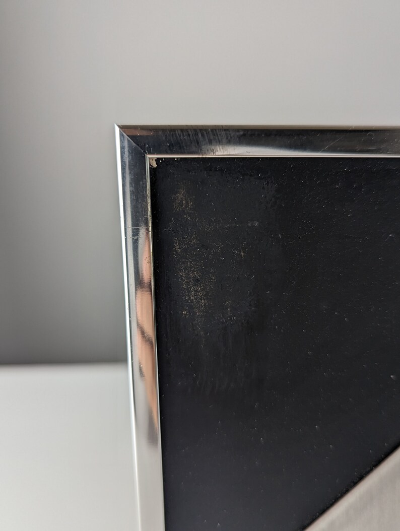 May include: Close-up of a silver metal picture frame with a black interior. The frame has a clean, modern design with a reflective surface. The black interior has a slightly textured appearance.
