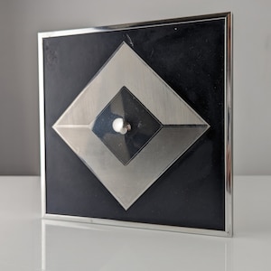 May include: Square black and silver decorative object with a geometric design. The object has a black background with a silver diamond shape in the center, and a smaller black diamond shape inside. A small silver ball is in the center.