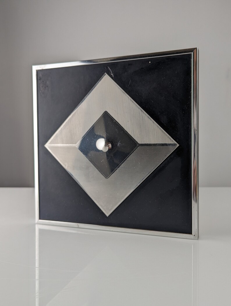 May include: Square black and silver decorative object with a geometric design. The object has a black background with a silver diamond shape in the center, and a smaller black diamond shape inside. A small silver ball is in the center.