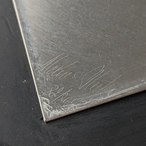 May include: A silver-colored, rectangular metal plate with etched cursive text and the numbers "22/8001". The plate has a brushed texture and is set against a black surface.